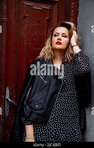 Image verticale de femme blonde moderne et souriante avec maquillage en veste et robe, debout près de la porte marron. Gros plan Banque D'Images