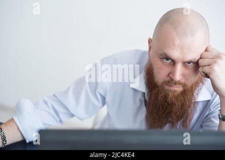 Un homme chauve focalisé avec une barbe rouge fixe avec attention à la caméra. Le Manager d'une chemise blanche est en colère contre le travail. Banque D'Images
