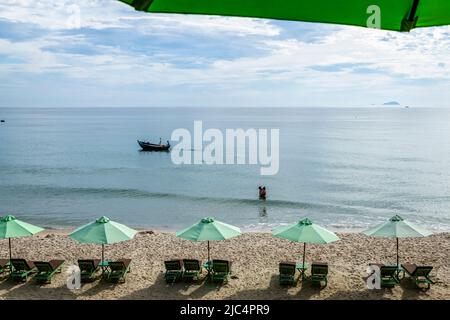 Une plage de Bang sur une matinée ensoleillée avec deux personnes dans l'eau et une rangée de chaises longues avec parasols bordent la plage de rivage. Banque D'Images