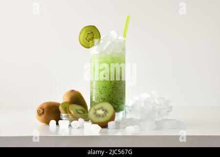 La neige fondue de kiwi boit dans un grand verre avec des fruits et de la glace pilée autour d'elle sur une table blanche avec un fond isolé. Vue avant. Composition horizontale. Banque D'Images