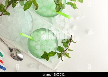 Verre avec boisson à la menthe glacée sur une assiette avec glace pilée et feuilles de menthe autour sur une table blanche. Vue de dessus. Composition horizontale. Banque D'Images