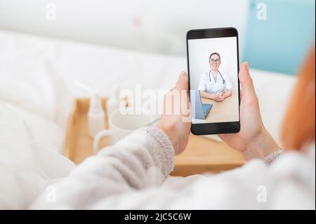 Le concept de traitement à distance. Vue arrière d'une femme au lit en pyjama avec un smartphone dans les mains. Un patient atteint de grippe regarde une vidéo médicale Banque D'Images