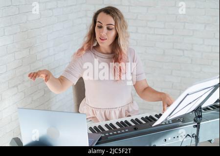 Une femme amicale joue le piano électronique et dirige un blog vidéo sur son ordinateur portable. Restez à la maison. Professeur d'instruments de musique. Musique d'apprentissage à distance Banque D'Images