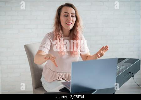 Une femme amicale joue le piano électronique et dirige un blog vidéo sur son ordinateur portable. Restez à la maison. Professeur d'instruments de musique. Musique d'apprentissage à distance Banque D'Images