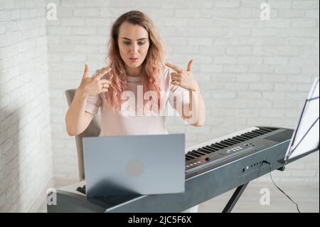 Une femme amicale joue le piano électronique et dirige un blog vidéo sur son ordinateur portable. Restez à la maison. Professeur d'instruments de musique. Musique d'apprentissage à distance Banque D'Images