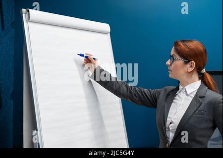 Une femme sympathique dans un costume d'affaires écrit sur un tableau blanc vierge avec marqueur. Une fille aux cheveux rouges fait une présentation au bureau. Belle femme Banque D'Images