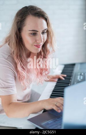 Une femme amicale joue le piano électronique et dirige un blog vidéo sur son ordinateur portable. Restez à la maison. Professeur d'instruments de musique. Musique d'apprentissage à distance Banque D'Images