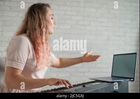 Une femme amicale joue le piano électronique et dirige un blog vidéo sur son ordinateur portable. Restez à la maison. Professeur d'instruments de musique. Musique d'apprentissage à distance Banque D'Images