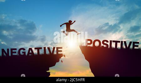 Silhouette homme sautant sur des mots négatifs et positifs sur les falaises avec ciel nuageux et lever de soleil. État d'esprit positif et concept de pensée positive. Banque D'Images