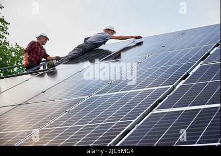 Les hommes qui travaillent à monter des modules solaires pour produire de l'électricité par l'effet photovoltaïque. Deux hommes techniciens solaires dans des casques de sécurité assemblant des panneaux solaires sous ciel bleu blanc. Banque D'Images