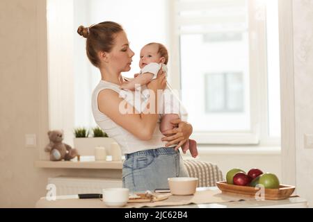 Jeune mère en Tshirt blanc parlant à son bébé tout en se tenant dans la cuisine à côté de la table avec de la nourriture Banque D'Images
