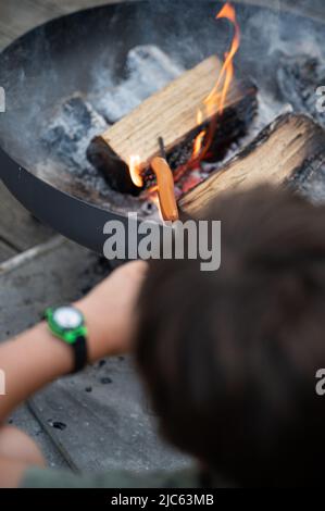 Vue sur la tête d'un garçon qui fait griller un hot dog sur un bâton au-dessus d'un feu ouvert dans un grill en fonte dans la cour intérieure. Banque D'Images