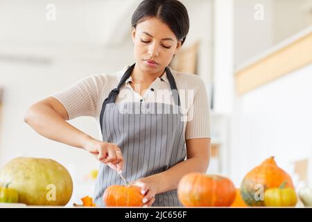 Jeune femme noire en tablier enlevant les graines de la citrouille tout en faisant le jack-o-lanterne à la maison Banque D'Images