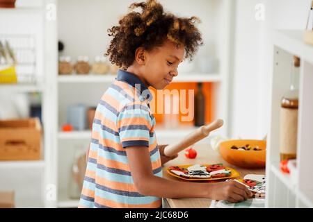 Mignon petit garçon afro-américain souriant dans un t-shirt coloré debout au comptoir de la cuisine et en utilisant une punaise tout en cuisinant des biscuits au pain d'épice Banque D'Images