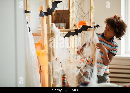 Grave mignon Noir garçon dans diable corne de serre-tête debout dans la salle de séjour et faisant la décoration de filet d'araignée pour Halloween Banque D'Images