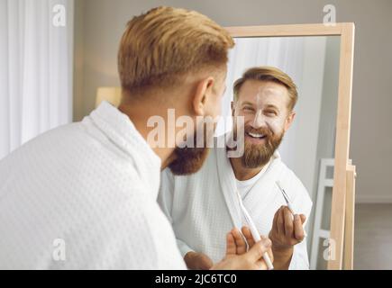 Heureux beau homme regardant dans le miroir, appliquant un masque blanc d'argile et souriant Banque D'Images