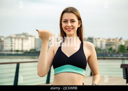 Heureuse femme sportive portant un soutien-gorge de sport debout sur le parc de la ville, en plein air souriant avec le visage heureux regardant la caméra et pointant vers le côté avec le TH Banque D'Images