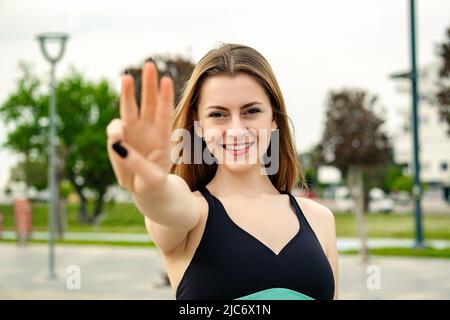 Heureuse femme sportive portant un soutien-gorge de sport debout sur le parc de la ville, à l'extérieur montrant et pointant vers le haut avec les doigts numéro trois tout en souriant confiant et Banque D'Images