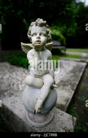 la petite figure d'ange endommagée envoie des voeux d'amour assis sur une boule de pierre à une tombe Banque D'Images