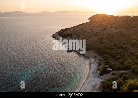 Voiture Offroad blanche avec tente sur le toit sur la côte grecque avec eau turquoise au premier plan et coucher de soleil en arrière-plan. Grèce. Banque D'Images