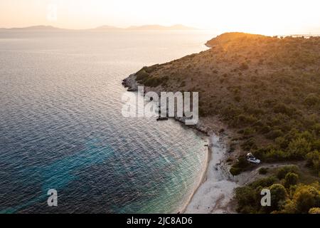 Voiture Offroad blanche avec tente sur le toit sur la côte grecque avec eau turquoise au premier plan et coucher de soleil en arrière-plan. Grèce. Banque D'Images