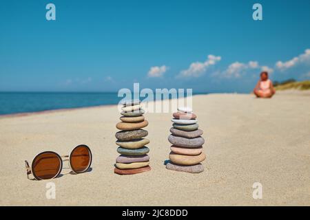 Cairn de pierres et de lunettes de soleil sur une plage de sable sur le fond d'une jeune femme assise dans une position de lotus Banque D'Images