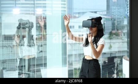 Jeune femme portant un casque de réalité virtuelle et interagit avec le cyberespace, se tenant près d'une grande fenêtre avec vue sur les bâtiments de la ville à l'extérieur Banque D'Images