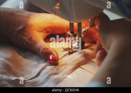 Photo des mains de femme avec vernis à ongles rouge en cours de couture robe de lin à l'aide de machine à coudre automatique à la maison Banque D'Images