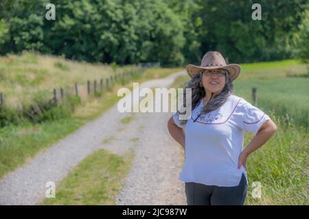 Femme mûre debout sur le chemin de campagne entre les terres agricoles néerlandaises, regardant la caméra, cheveux gris ondulés troués par le vent, chapeau de cowboy, arbres luxuriants dans le bac flou Banque D'Images