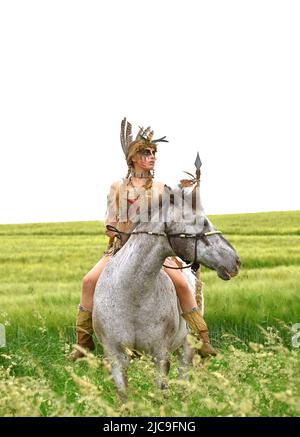On voit une jeune fille indienne monter son poney dans la prarie. Elle est habillée comme indienne amérindienne et tient une lance dans sa main. Banque D'Images