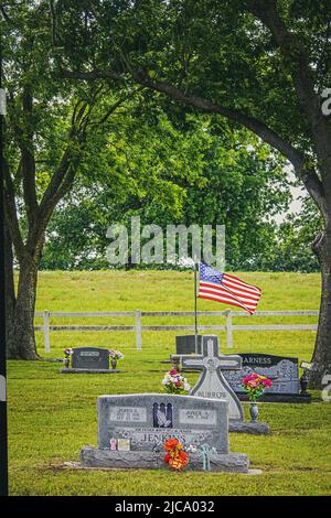 2019 05 25 Mannford OK cimetière de pays des États-Unis avec des pierres tombales et des fleurs religieuses et un drapeau américain entre deux arbres avec une clôture blanche et m Banque D'Images