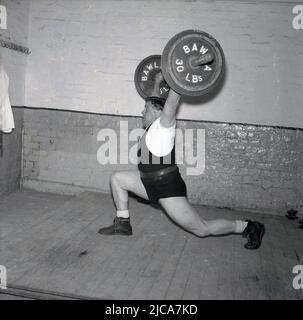 1957, historique, haltérophilie, à l'intérieur d'une salle de gym sur une planche en bois, un entraînement d'haltérophilie mâle, levant la barbell faisant le propre-et-jerk, Stockport, Manchester, Angleterre, Royaume-Uni. Sur la barbell est 60lbs. Le Clean-and-jerk est l'un des deux ascenseurs de l'haltérophilie de compétition ou de style olympique. Il porte des chaussures d'haltérophilie et la ceinture d'haltérophilie traditionnelle en cuir. Sur la barbell, un logo avec les initiales BAWLA, la British amateur Weightlifting Association, nommée ainsi en 1911 après avoir été fondée en 1910 comme la BWLA. Banque D'Images