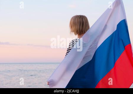 Vue arrière blonde femme agitant le drapeau national de la Russie dehors mer coucher de soleil l'été - drapeau russe, pays, patriotisme, jour de la Russie et le 12 juin. Banque D'Images