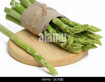 Un bouquet d'asperges fraîches enveloppées de papier brun se trouve dans une assiette en bois sur fond blanc Banque D'Images