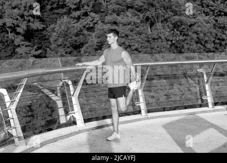 Homme sportif en vêtements de sport saisissez la cheville avec la main étirant la jambe après l'entraînement en plein air, étirez Banque D'Images