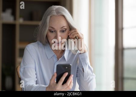 Surprise femme âgée regardant à l'écran de téléphone portable lire des nouvelles fantastiques Banque D'Images