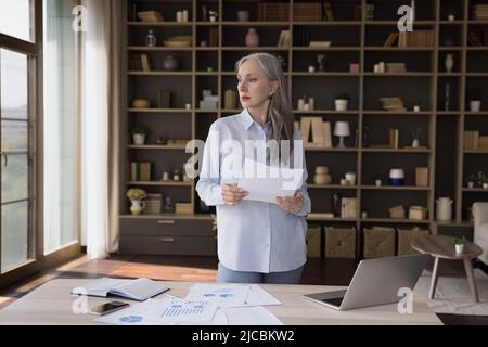 Une femme d'affaires sérieuse et âgée distrait de la paperasserie en regardant la fenêtre Banque D'Images