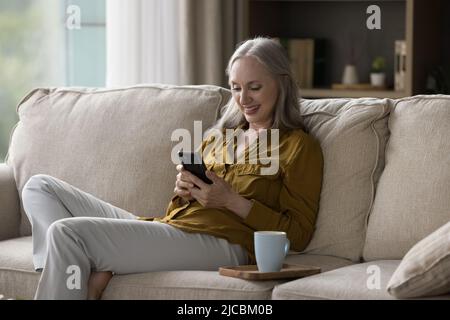 Bonne femme âgée se reposant sur un canapé avec un smartphone Banque D'Images