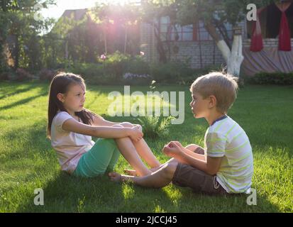 En été, garçon et fille de 6-7 ans s'assoient sur l'herbe dans l'arrière-cour du soleil couchant dans le jardin. Parler et jouer dans le parc vert ensoleillé. Réfrigérateur Banque D'Images