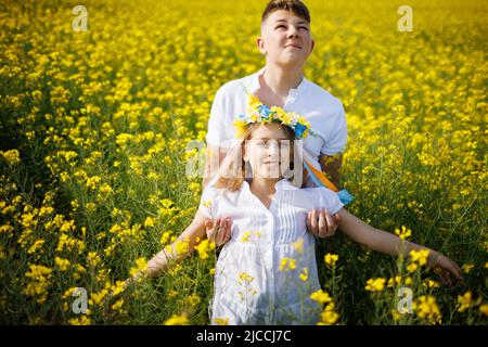 Grand frère adolescent fort attrape sa petite sœur joyeuse et joyeuse et sa couronne de fleurs ukrainiennes qui tombe entre ses mains, en vous de colza Banque D'Images