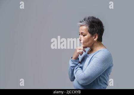 Portrait de mûr réfléchi cheveux gris femme d'affaires posant avec les mains pliées touchant le menton et de copier l'espace et de placer pour le produit sur la gauche isolé sur fond blanc. Concept de beauté vieilli. Banque D'Images