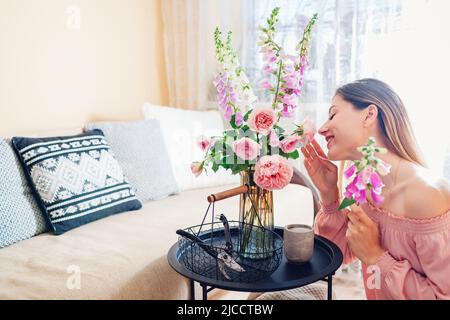 La femme sent des fleurs en agençant le bouquet avec des roses et des foxgants à la maison. Fleurs fraîches mises dans vase sur table. Décoration intérieure et estivale Banque D'Images