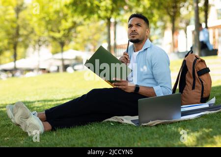 Concept de formation en ligne. Étudiant afro-américain attentif étudiant en plein air, en utilisant un ordinateur portable et en prenant des notes Banque D'Images