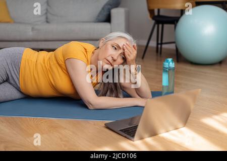 Femme séchée sur un tapis de yoga devant un ordinateur portable et essuyant son front, prenant une pause après l'entraînement à la maison Banque D'Images
