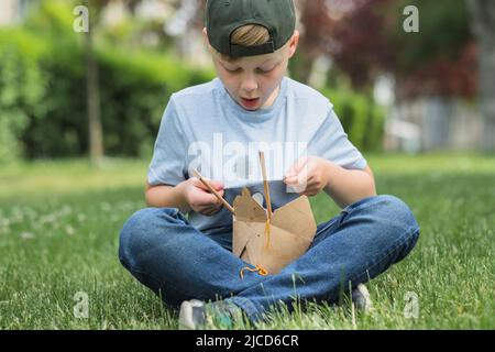 Le garçon montrant une sale tache de sauce sur les vêtements. Enfant mangeant de la nourriture à emporter assis sur l'herbe dans le parc. À l'extérieur Banque D'Images