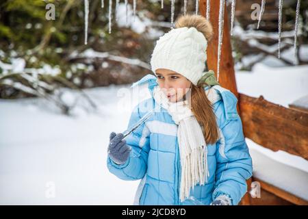 Bien pensé gai petite fille en hiver chaud vêtements lumineux drôle licks un froid vif transparent icicle tout en se tenant dans une forêt de pins enneigés Banque D'Images