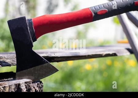 Une nouvelle hache moderne avec une poignée rouge colle d'une souche en bois contre un pré vert lors d'une journée ensoleillée d'été. La hache s'est coincée dans la souche. Lame AX vers l'intérieur Banque D'Images