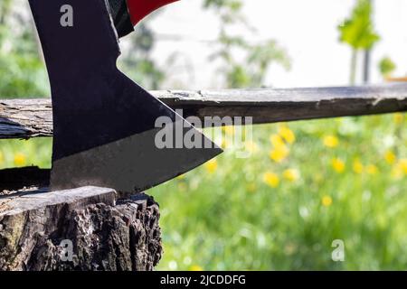 Une nouvelle hache moderne avec une poignée rouge colle d'une souche en bois contre un pré vert lors d'une journée ensoleillée d'été. La hache s'est coincée dans la souche. Lame AX vers l'intérieur Banque D'Images