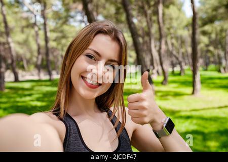 Bonne femme brune portant un soutien-gorge de sport noir debout sur le parc de la ville, à l'extérieur montrant le pouce et prenant des photos de selfie. Regarder l'appareil photo. Banque D'Images