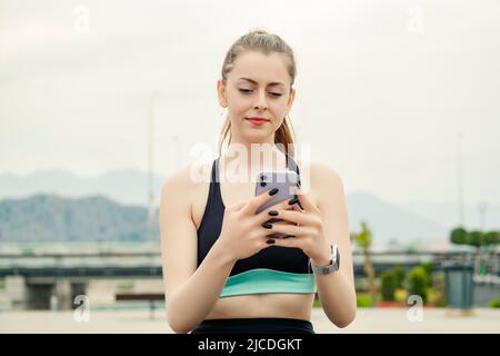 Jeune femme brune portant des vêtements de sport sur le parc de la ville, à l'extérieur regardant l'écran du téléphone et utilisant le téléphone. Envoyez des messages avec vos amis, regardez des vidéos Banque D'Images